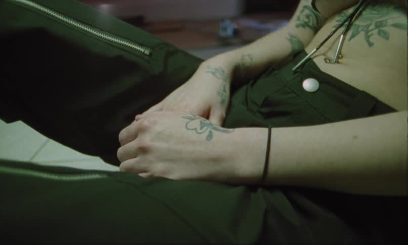 A tattooed person sits on the floor while light flashes in dark pants with zippers one hand resting near a white button and a black hair tie on the wrist