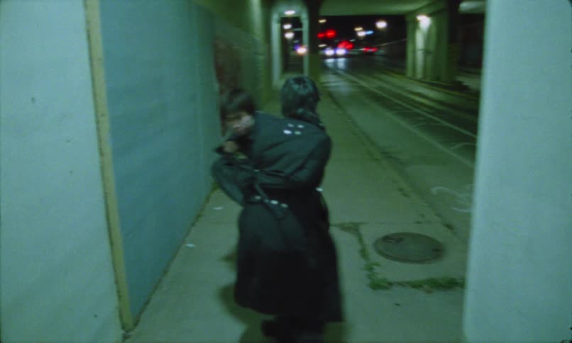 Two people both dressed in dark clothing rapidly moving away from the camera down a narrow graffiti marked walkway and runs alongside an active road under a concrete overpass or bridge with the lights of a passing vehicle