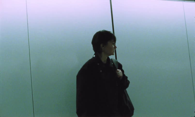 A young man wearing a black jacket and carrying a backpack looks frustrated