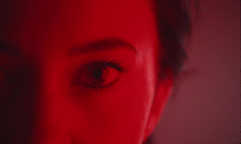 A young woman inside a room illuminated by red light looks slightly angry