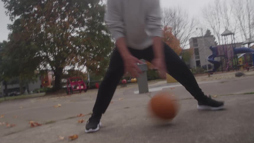 A person in a gray sweatshirt dribbles a basketball low at a park playground