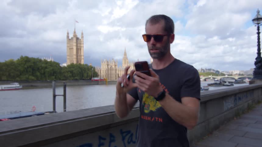 A young man walking on a bridge at midday wearing sunglasses and a branded tshirt holds his phone to his ear to make a call