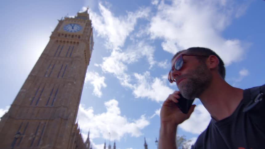 A bearded man wearing sunglasses is filmed from a low angle against a bright blue cloudy sky as he holds a smartphone to his ear and talks while standing near the base of a tall clock tower and a bus passing in the background