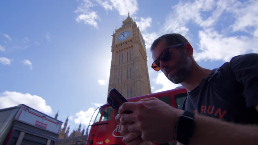 A young man wearing sunglasses checks his watch then continues typing on his mobile phone while standing on the sidewalk as a doubledecker bus passes by with a clock tower in the city background at midday