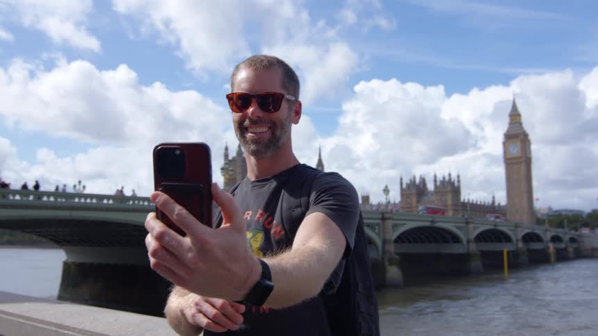 A man in sunglasses holds his phone up to take a selfie at a landmark