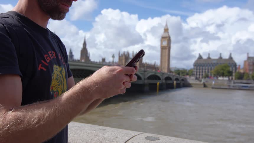 The video features a closeup of a man on the phone wearing sunglasses and a black tshirt standing outdoors on a sunny dayhe ends his call and then looks down to interact with his phone with the iconic big ben clock tower and the houses of parliame