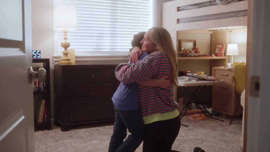 Mother hugs son while kneeling in a bedroom