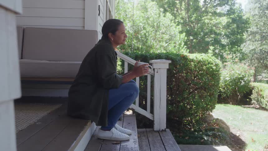 A woman sits on a porch holding a coffee mug and gazing at the greenery