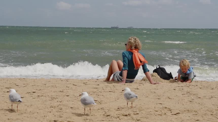 Woman and child sit in the sand as birds watch