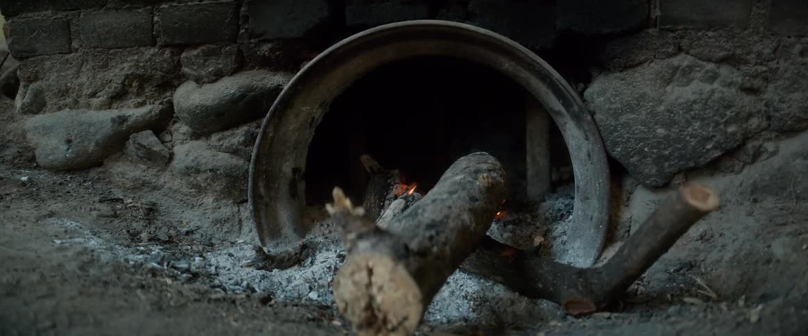 The video displays a closeup shot of a rustic outdoor stone and brick oven or fireplace showing the opening covered by an inverted metal vessel or bowltwo wooden logs are positioned across the opening and ash covers the ground around it with small