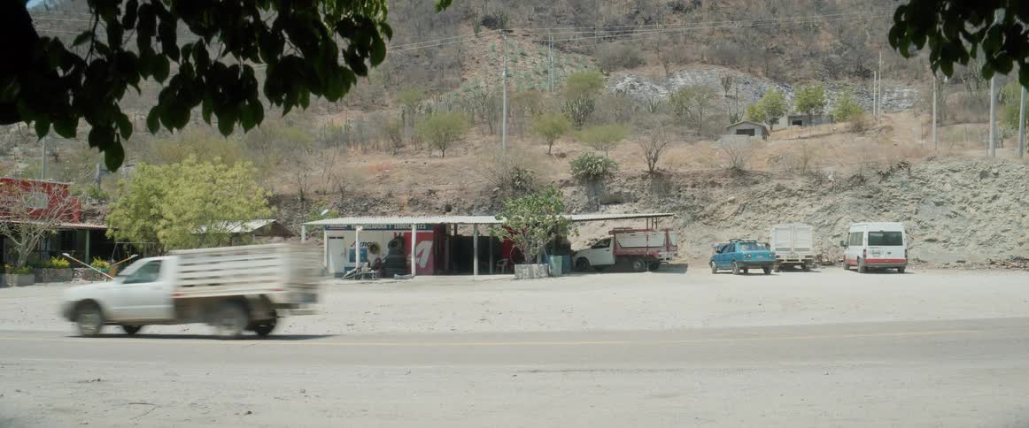 A roadside garage with several parked vehicles sits below a dry hilly landscape framed by overhanging leaves