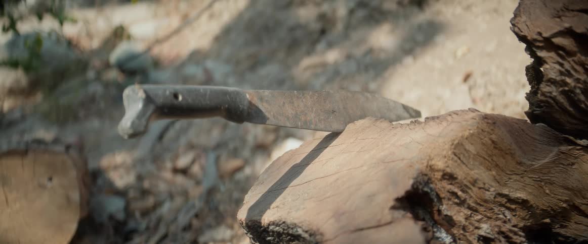 A closeup shot orbits a large rustic knife resting on a rough piece of wood its worn blade catching the bright outdoor sunlight