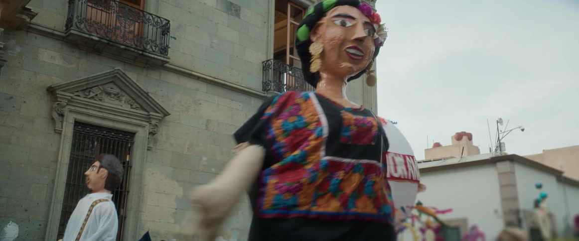A towering papiermch figure in a colorful embroidered dress moves through a city street parade