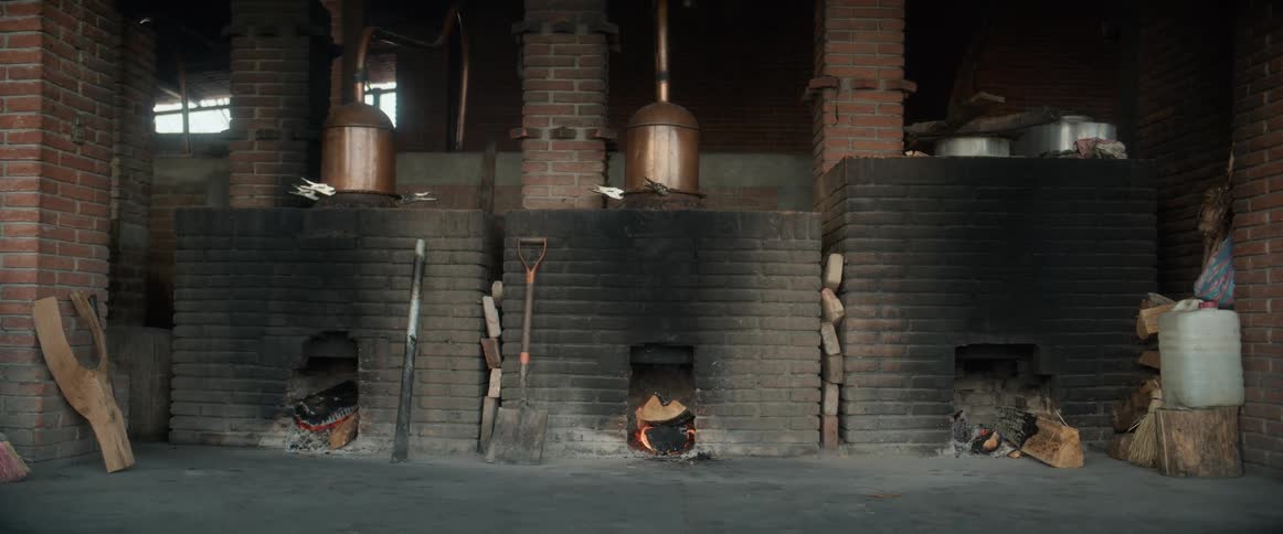 Three brick furnaces are built into a long dark brick wall with glowing fires visible inside the arched openings featuring copper pot stills and tall brick chimneys above them