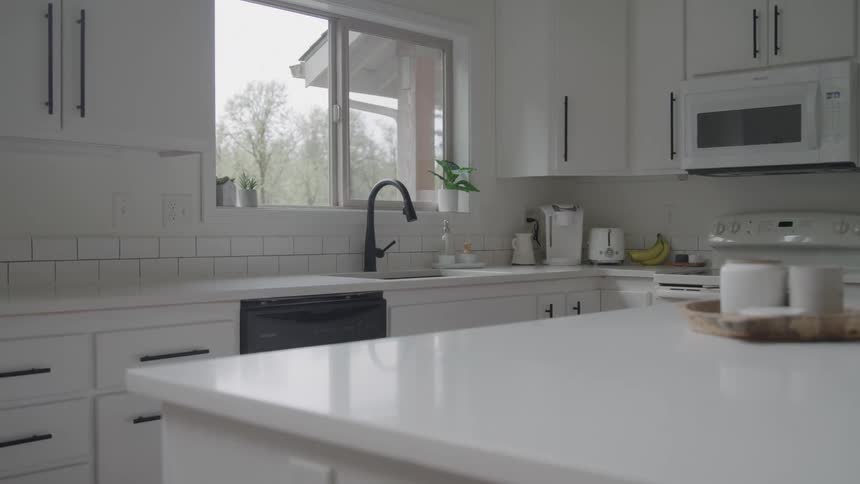 Modern kitchen dominated by white cabinets and quartz countertops featuring a black sink faucet centered under a window that reveals a blurred overcast outdoor view of trees
