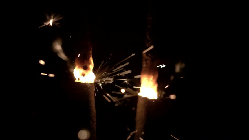 A closeup shot focuses on the glowing burning ends of two sparklers in nearcomplete darkness with bright yellowwhite sparks rapidly shooting outwards and falling away from the fiery wicks