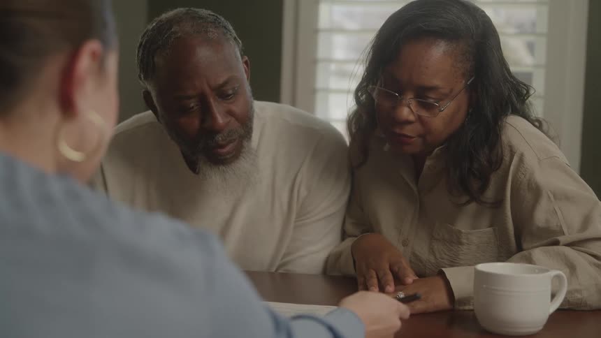 Two older black adults are listening to another woman talking and showing them a piece of paper