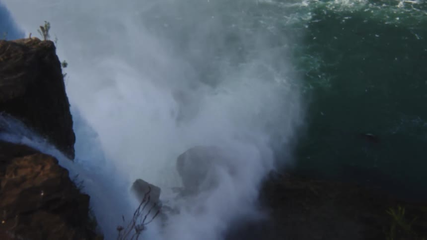 River water flowing powerfully over the rocky precipice of a large waterfall where a thick cloud of mist is generated below and a small faint rainbow arc is visible over the spray