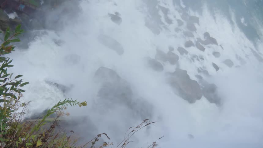 A misty cascade crashes over scattered rocks while grasses in the foreground bend toward the roaring water