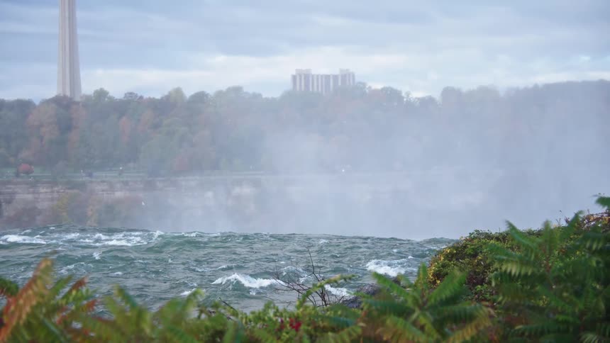 Mist rises above a powerful waterfall surrounded by greenery and distant buildings