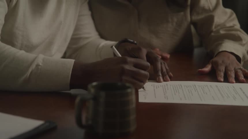 An elderly couple is seated across a wooden table from an unseen person as they review and sign a document culminating in a moment of shared joy and a celebratory hug