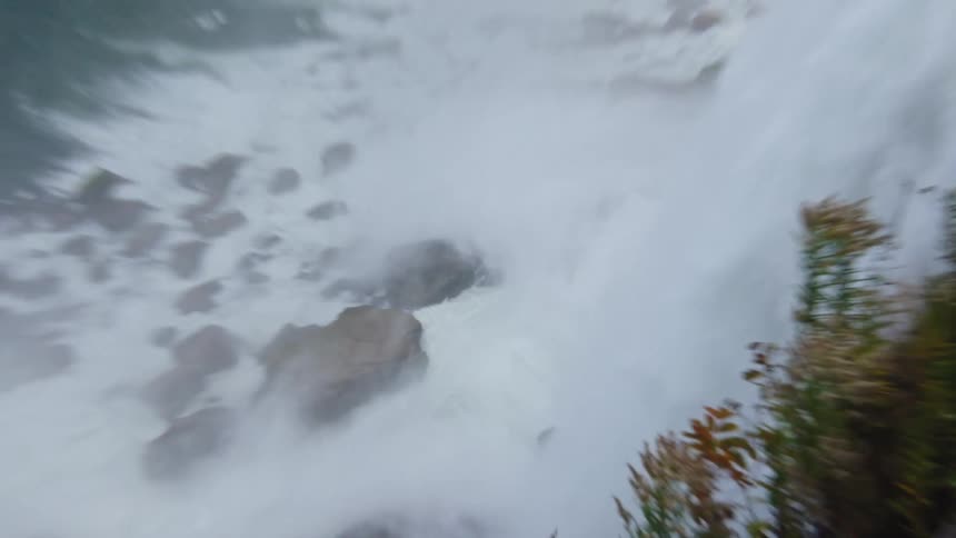 A powerful waterfall cascades down a rocky cliff face generating a vast cloud of white mist that obscures the view of the churning water below while in the immediate foreground a few sprigs of brown and green vegetation cling to the wet bank