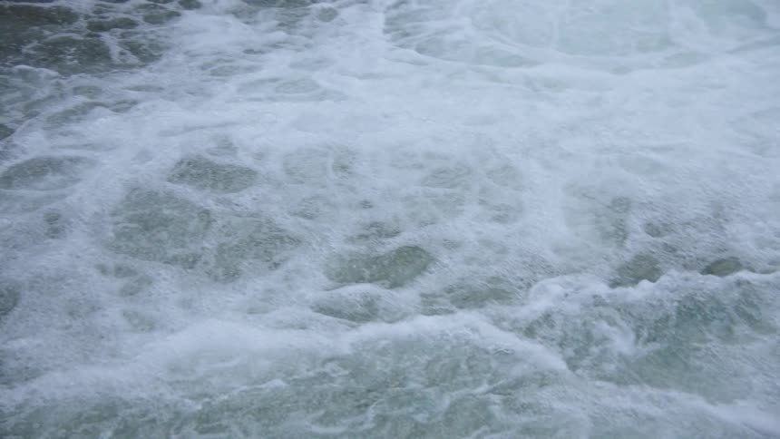 The violent agitation of a large body of water completely covered in thick churning white foam which gradually subsides to reveal the darker turbulent surface of the rushing current where a lone piece of wood sticks up from the swirling depths
