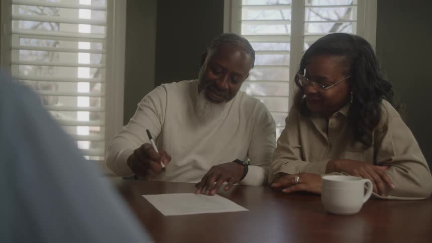 A man and woman sit with an advisor who points to a document on the table the couple hug after the man signs