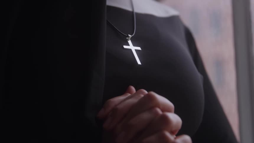 A closeup of a nun clasping her hands in prayer indoors