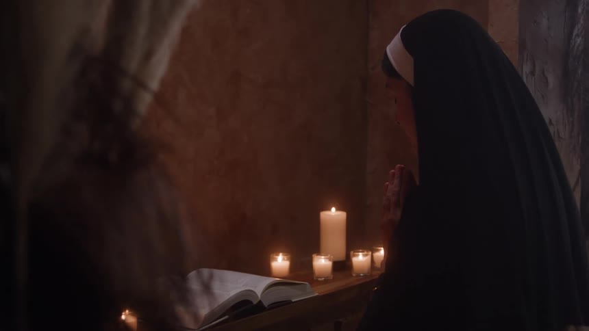A nun prays beside a lit candle and open book in a dimly lit room