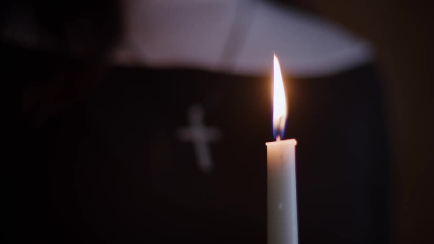 A closeup of a nun holding a candle while praying in a dimly lit room