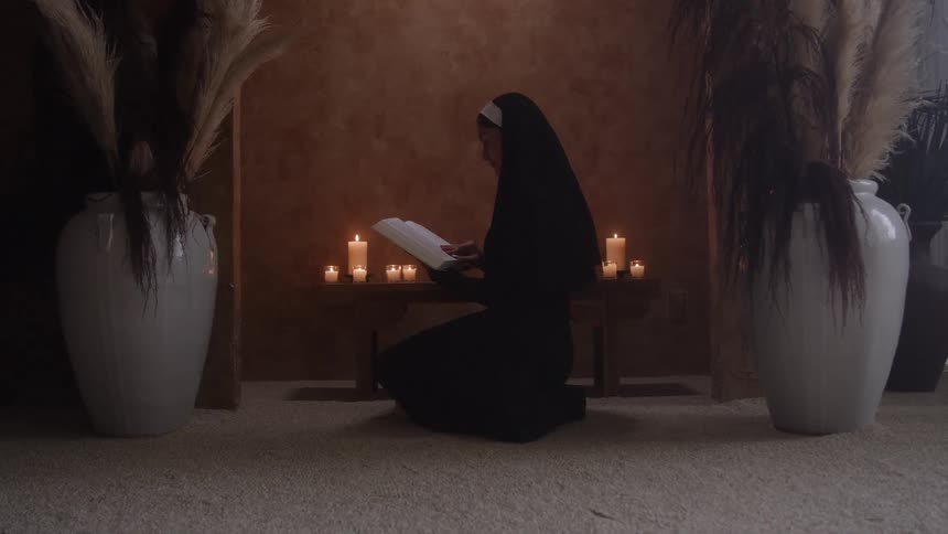 A nun in a black nuns habit kneels on a lightcolored floor intently reading an open book illuminated solely by the warm glow of several small lit candles placed on a low table in front of her flanked by two large white vases holding dried grass
