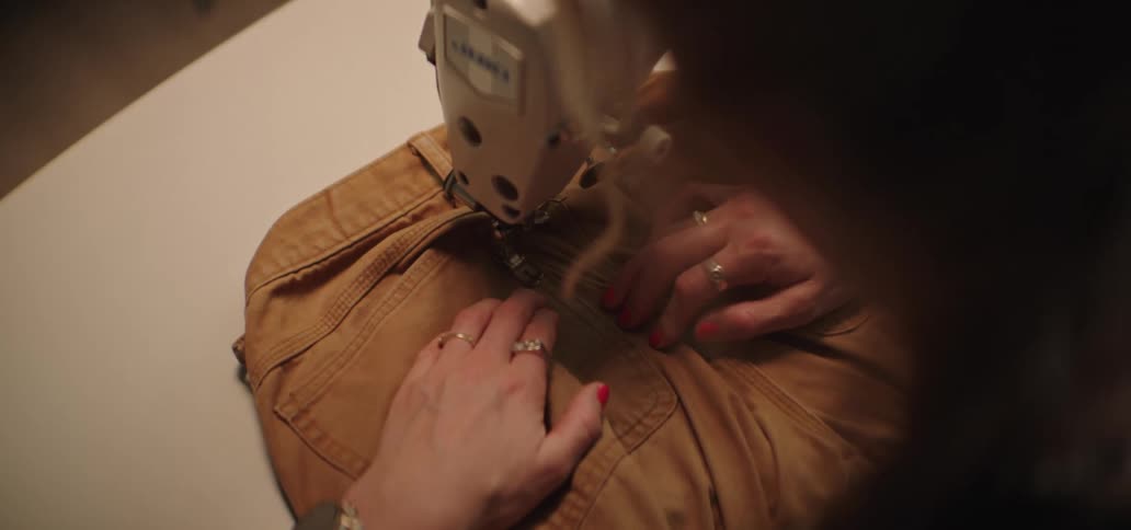 The video provides a closeup overtheshoulder view of a person using an industrial sewing machine to stitch a piece of tan or brown denimcanvas fabric likely a jacket or bag their hands adorned with rings and red nail polish expertly guide the mat