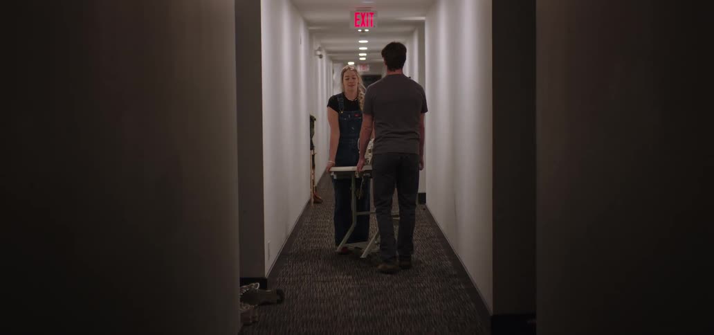 The video shows a man and a woman in a dimly lit long hallway likely in an apartment building or hotel walking towards the camerathe woman is pushing a luggage cart or moving dolly in front of her and an illuminated red exit sign is visible at t