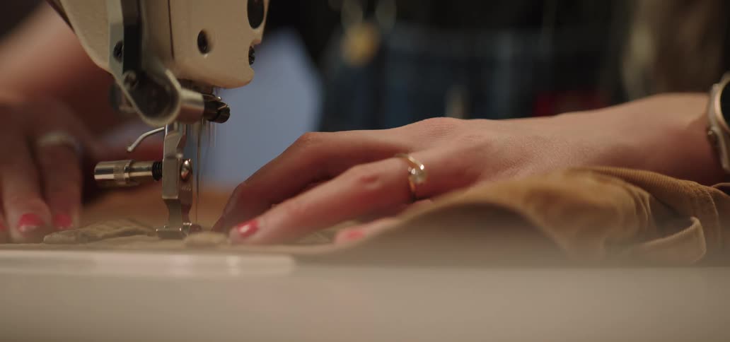 A close view of a sewing machine shows the hands of a woman with painted fingernails feeding brown fabric under the needle to sew a seam in a brightly lit tailor shop