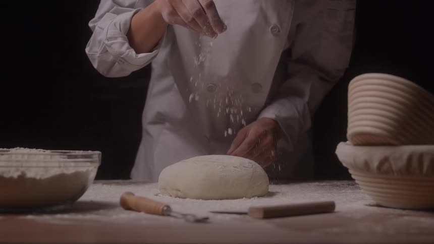 A baker sprinkles flour over a round dough on a wooden surface