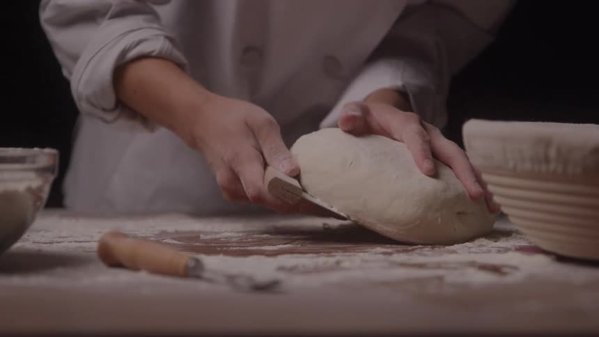 A baker uses a scraper to pick up a dough loaf and put it in a bowl