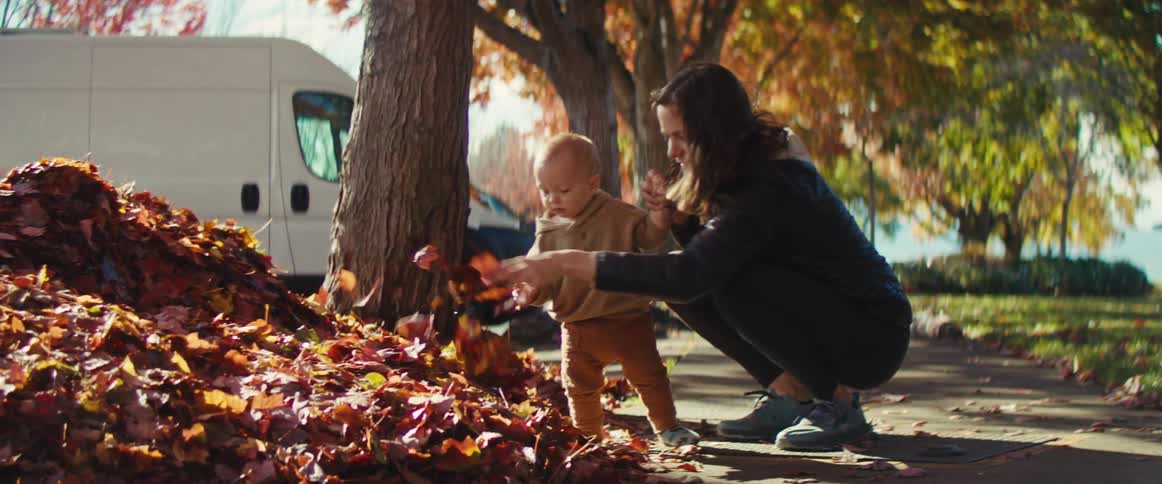A young mother and her male toddler play with fallen leaves on the sidewalk during daytime