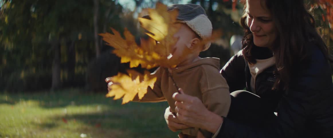 A mother shows a toddler maple leaves outdoors and the child playfully grabs at them