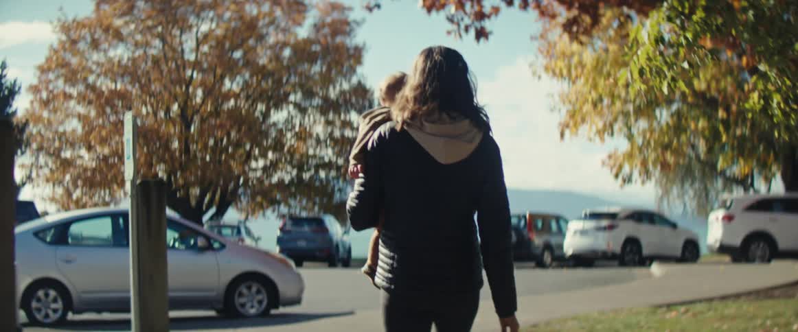 Woman walks carrying a child in a treelined parking area during autumn