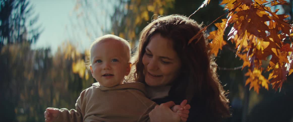 Mother holds her smiling child as they stand in a field near the trees on an autumn day