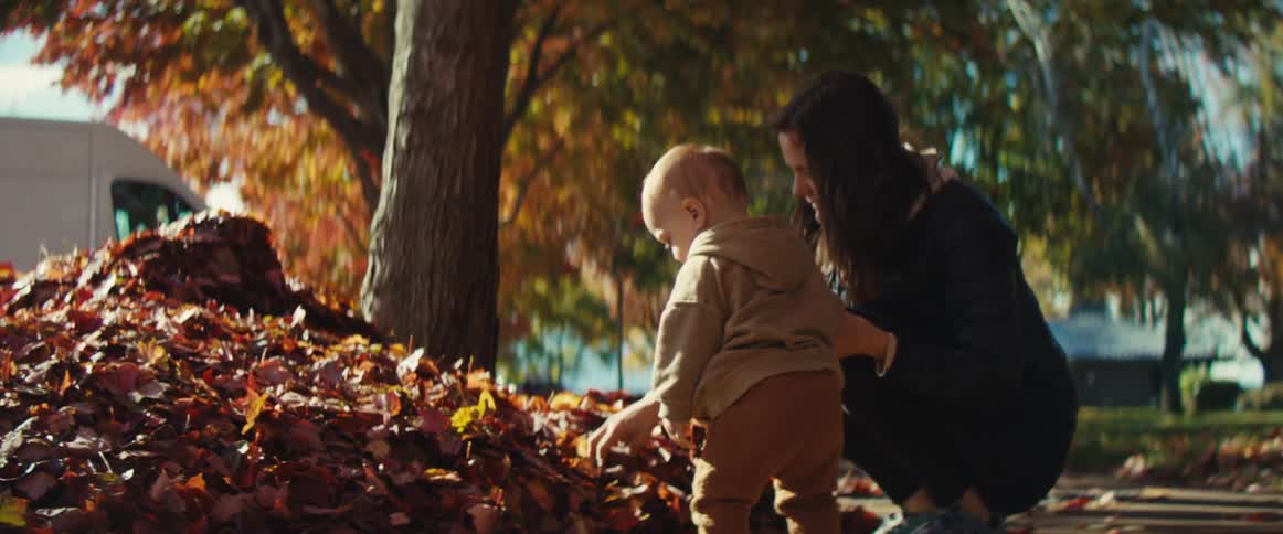 A woman crouching down holding the hand of a little boy who is playing with a pile of dry leaves
