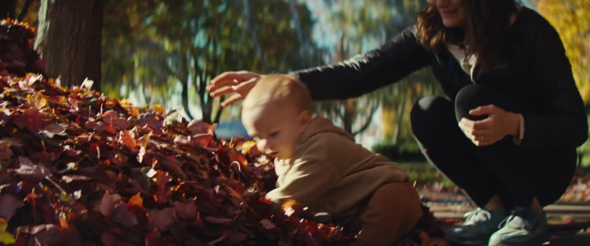 The video shows a baby or toddler dressed in a tan outfit standing near a large pile of bright red and orange autumn leaves outdoors on a sunny daya woman presumably the mother kneels beside the child gently encouraging them to interact with the lea