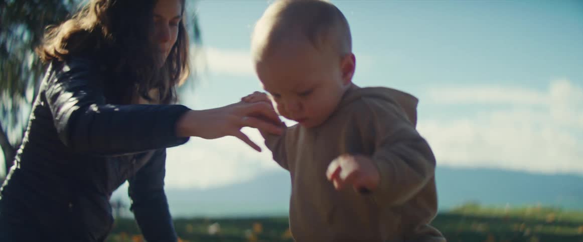 A mother kneels down holding the hands of her baby who is clad in a tan outfit as the child attempts to take tentative steps outdoors on a grassy sunlit field