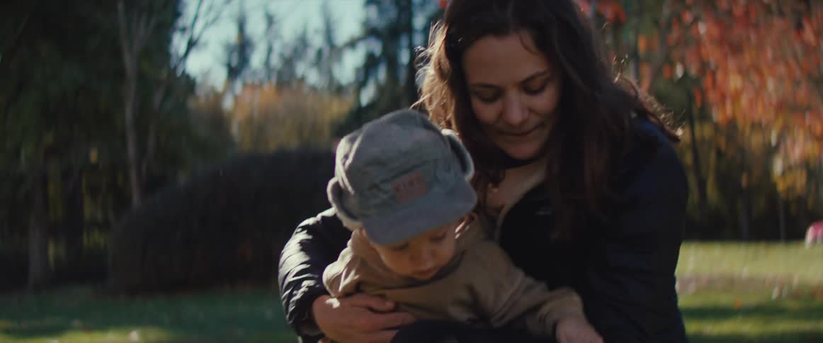 A woman crouches with a baby in her arms as she gently offers a bright yellow leaf branch to the child who leans in curiously