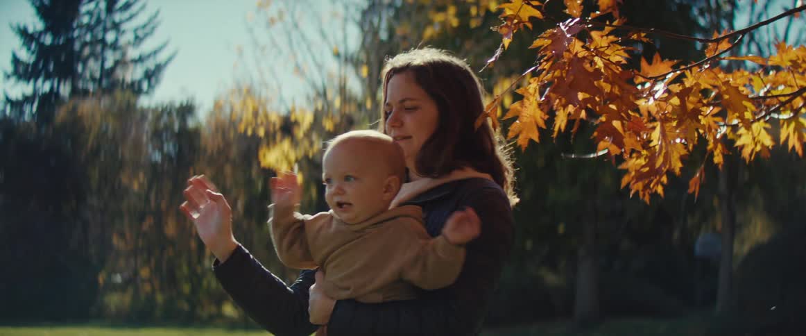 A young mother carrying her playful baby while standing outdoors in a park during daytime