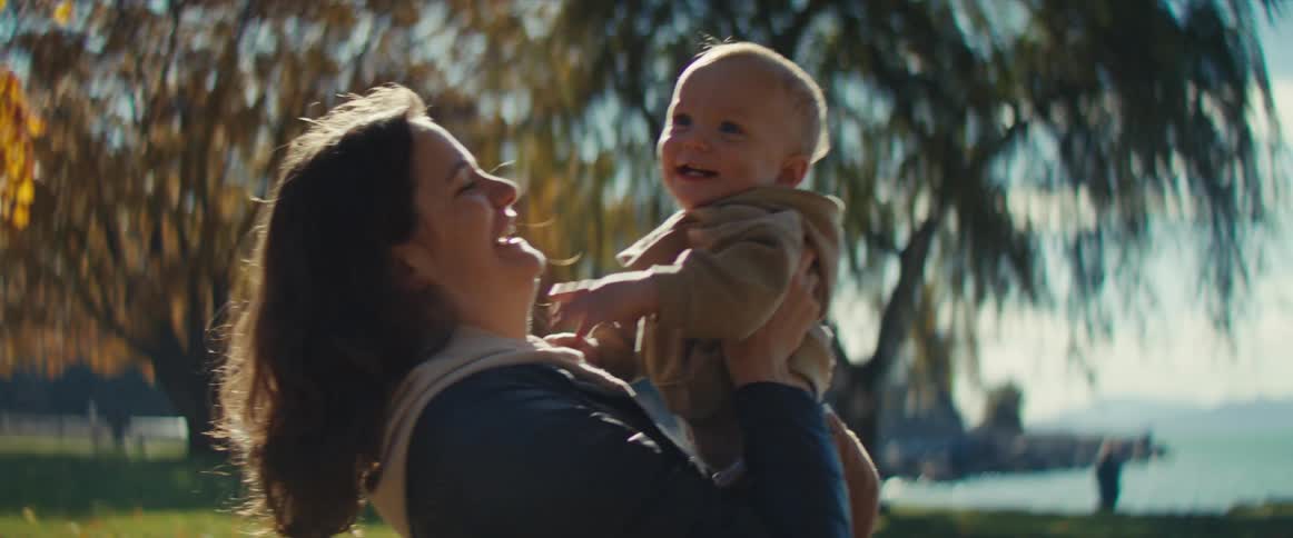 A woman is outdoors in a sunny park setting holding a smiling baby in her armsshe laughs and kisses the babys cheek as they look at each other with the sun backlighting them beneath tree foliage