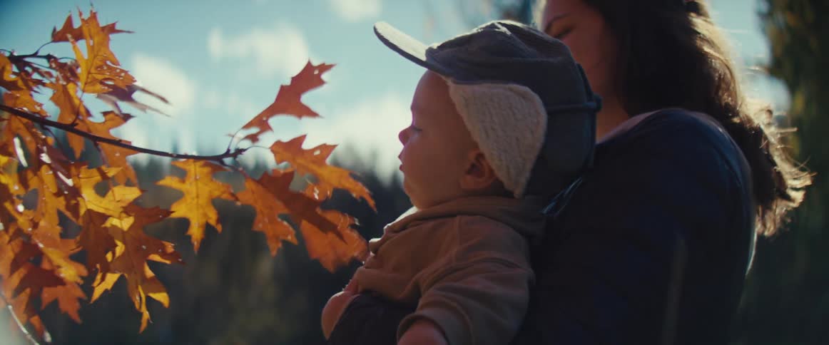 The video is a closeup shot of a mother holding a baby or young toddler who is wearing a soft cap with ear flaps outdoors during the autumnthe child reaches out to touch and play with the bright orange and red leaves of a branch in the foreground il
