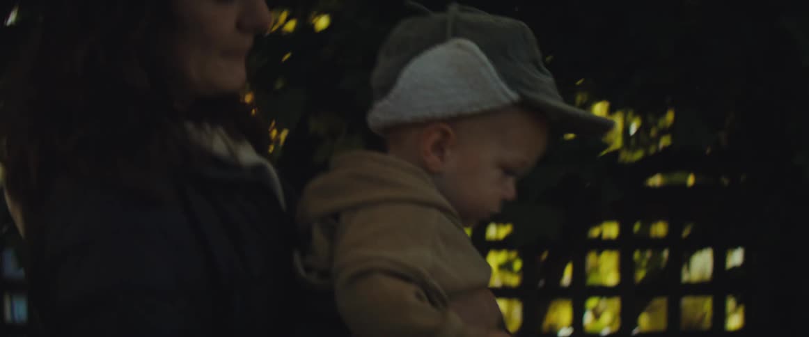A mother carrying her son who is wearing a cap on an outdoor walk near some trees