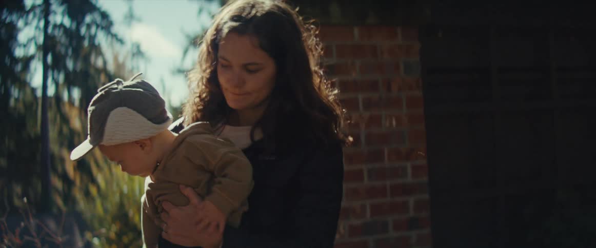 A mother picks up her baby from the ground outdoors with the baby wearing a hat on a sunny evening